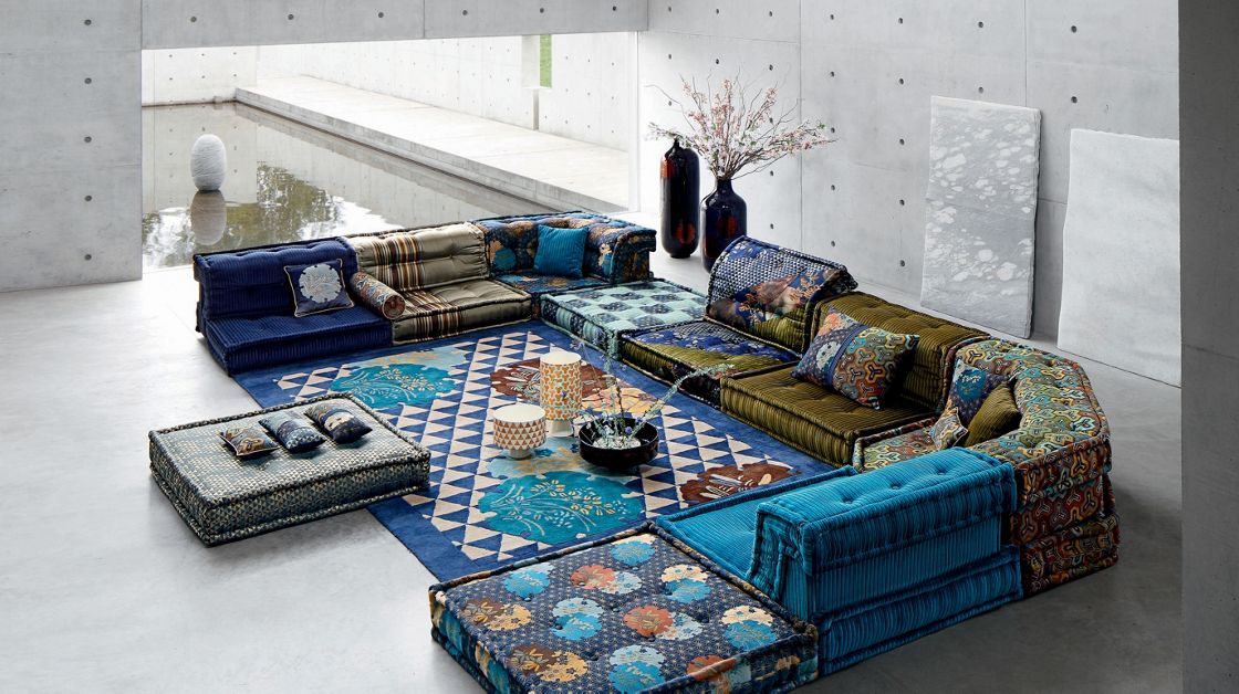 Canapé modulaire Mah Jong de Roche Bobois, issu d’une collaboration avec le styliste Kenzo Takada. Composé de coussins aux motifs et textures variés inspirés de l’art textile japonais, ce canapé bas repose sur un tapis graphique bleu et blanc dans un intérieur moderne en béton avec vue sur un bassin d’eau.