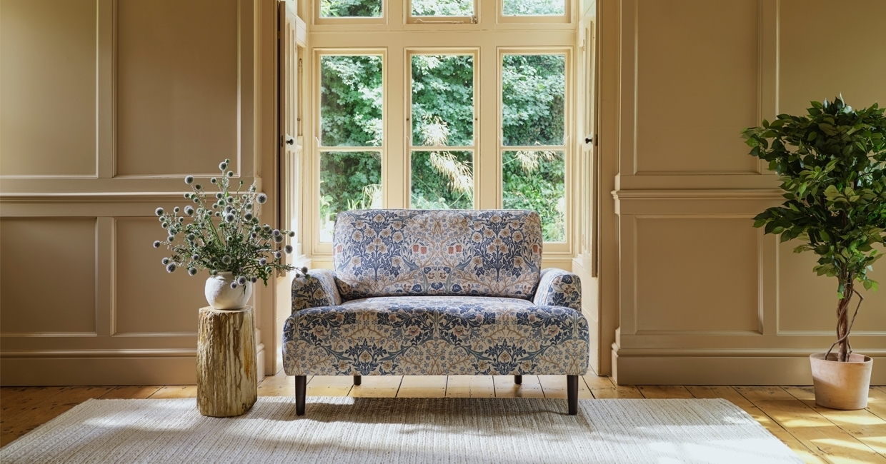 Canapé deux places issu de la collaboration entre Swyft Home et Morris & Co, aux motifs floraux bleu et rose, placé dans un salon élégant avec boiseries murales beiges, grandes fenêtres à carreaux et décoration végétale.