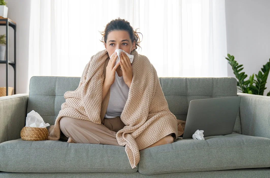 Femme assise sur un canapé, emmitouflée dans une couverture, en train de se moucher à cause des allergies.
