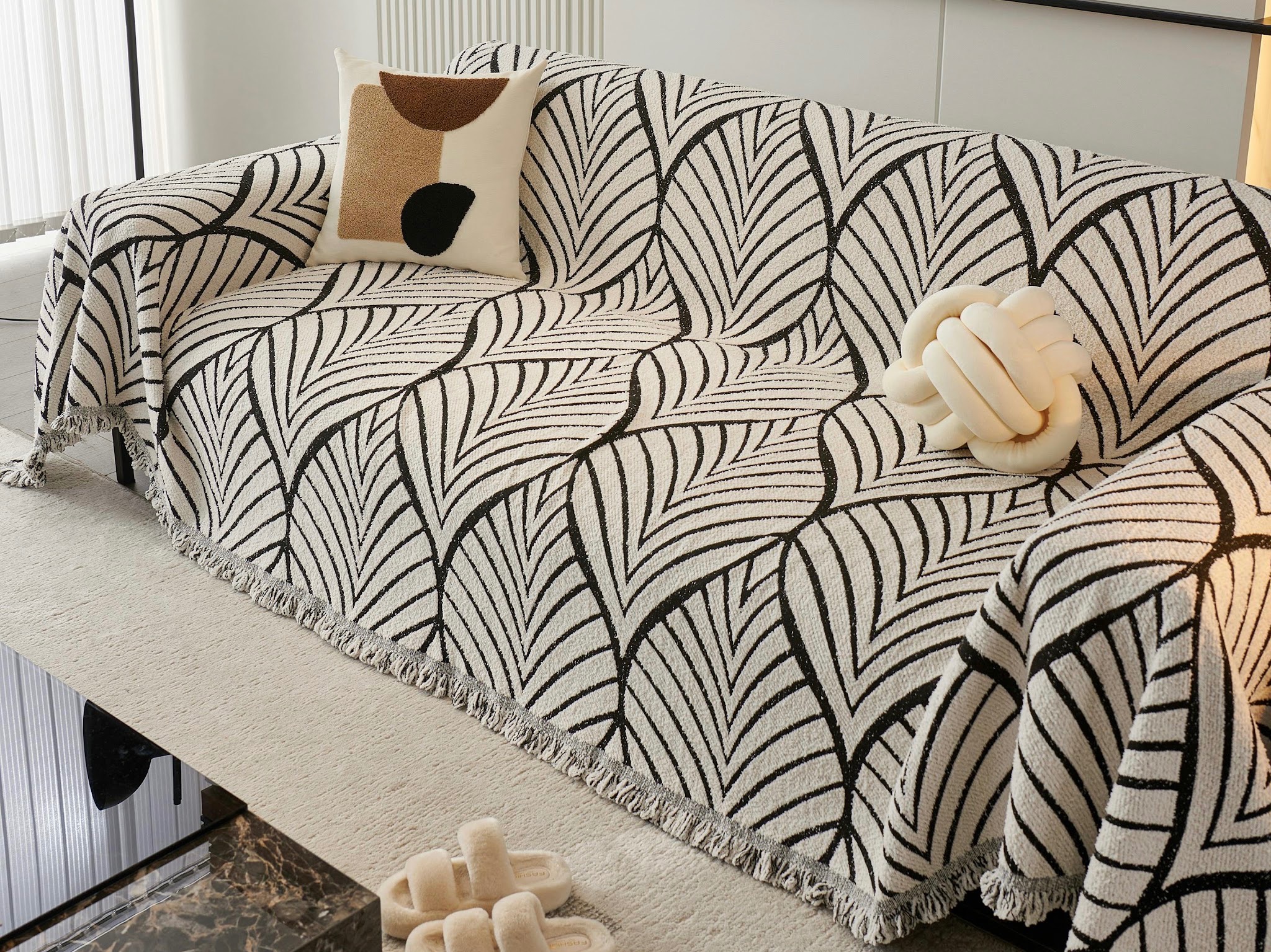 Un canapé élégamment recouvert d’un jeté noir et blanc à motifs de feuilles, avec un coussin noué et une paire de chaussons moelleux posés au sol.