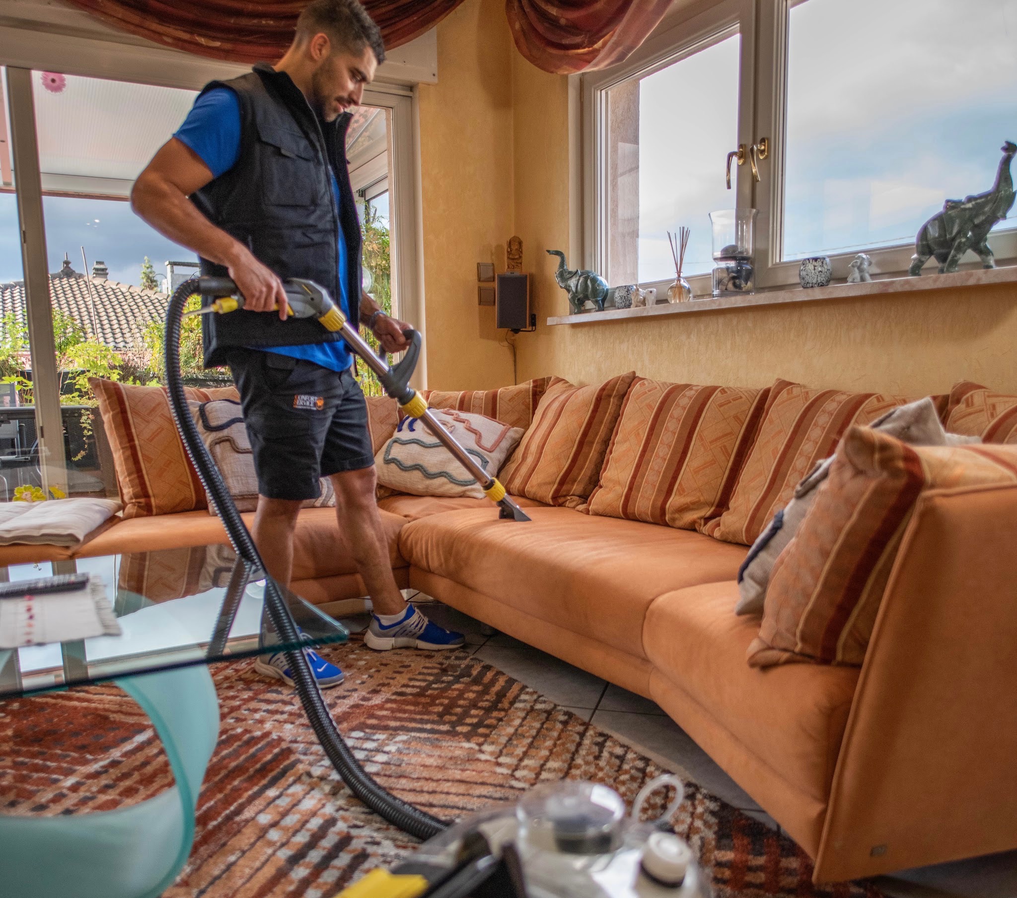 Un technicien de Confort Service en gilet de travail nettoie en profondeur un canapé d’angle orange à domicile avec un aspirateur extracteur, dans un salon.