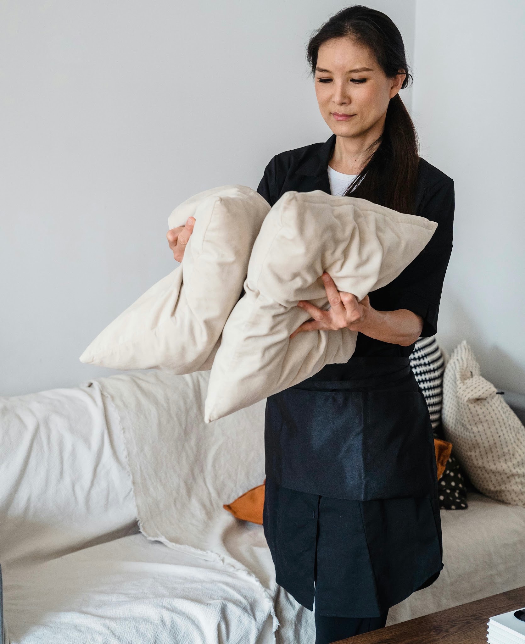 Une femme en tablier noir arrange soigneusement des coussins beige dans une pièce lumineuse et épurée, avec le soin