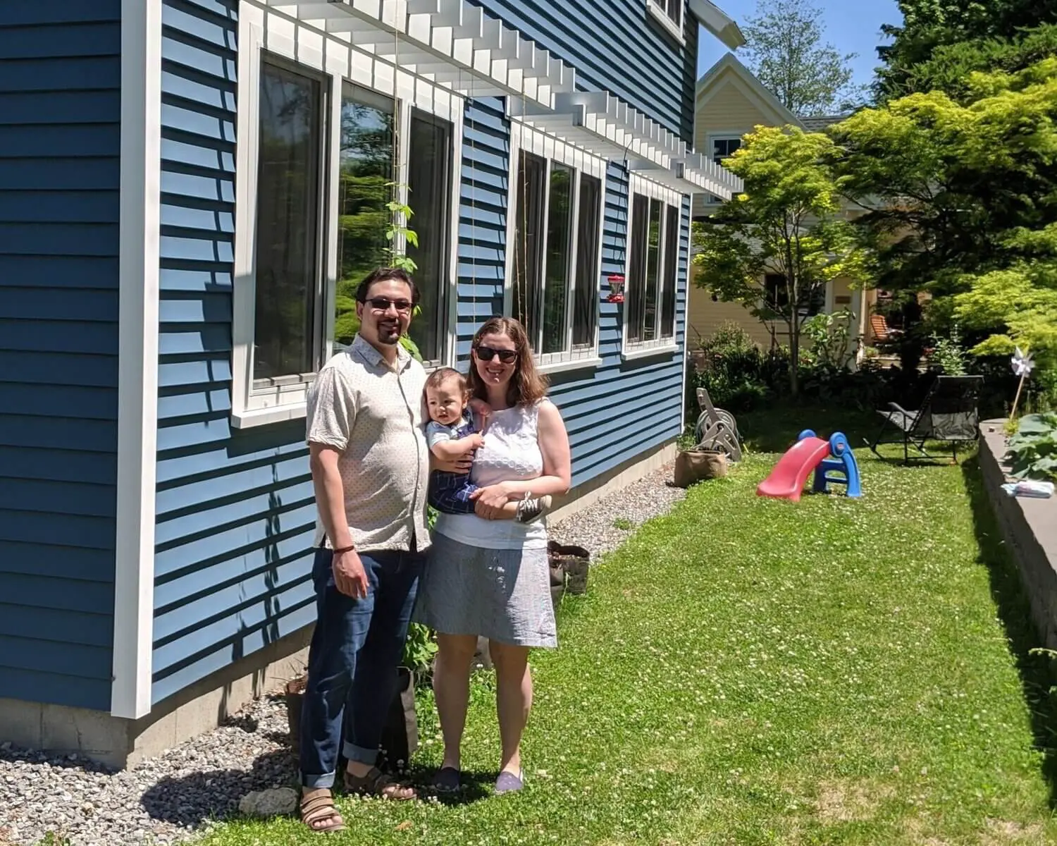 Family of three standing in sunny backyard next to blue house with slide