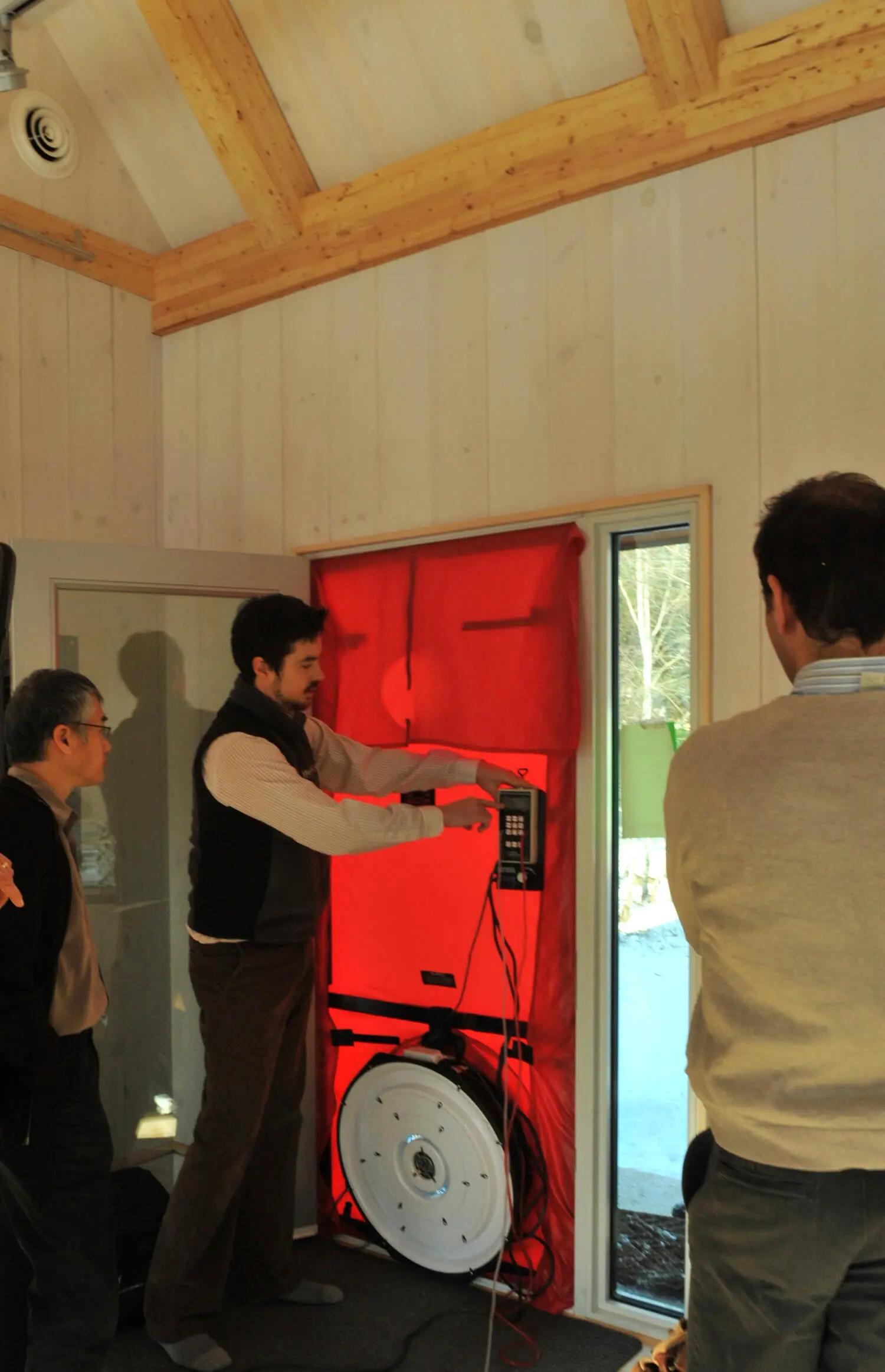 Technicians examining a red blower door setup in a wooden-paneled room