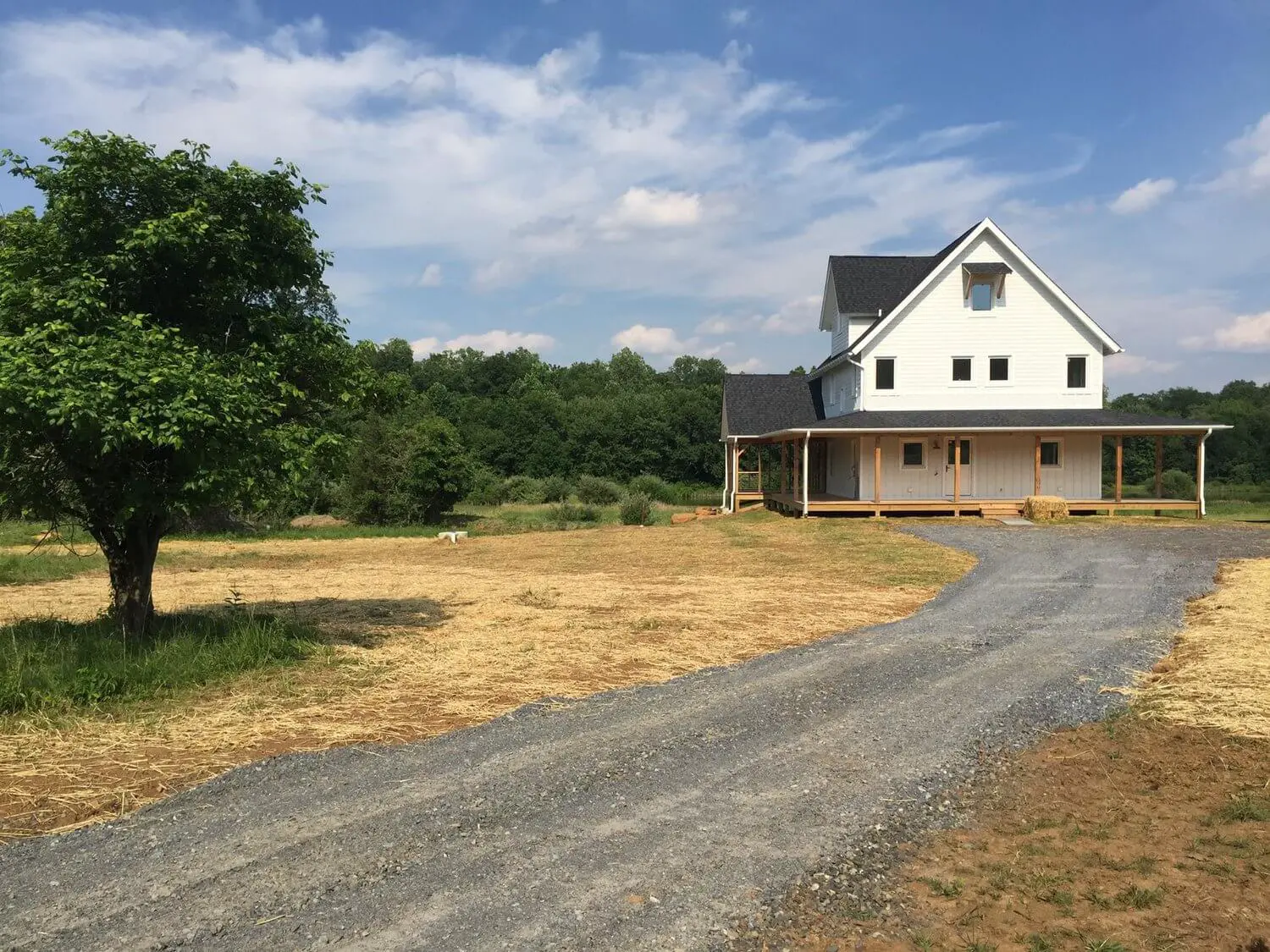 White farmhouse with wraparound porch on gravel driveway near green tree