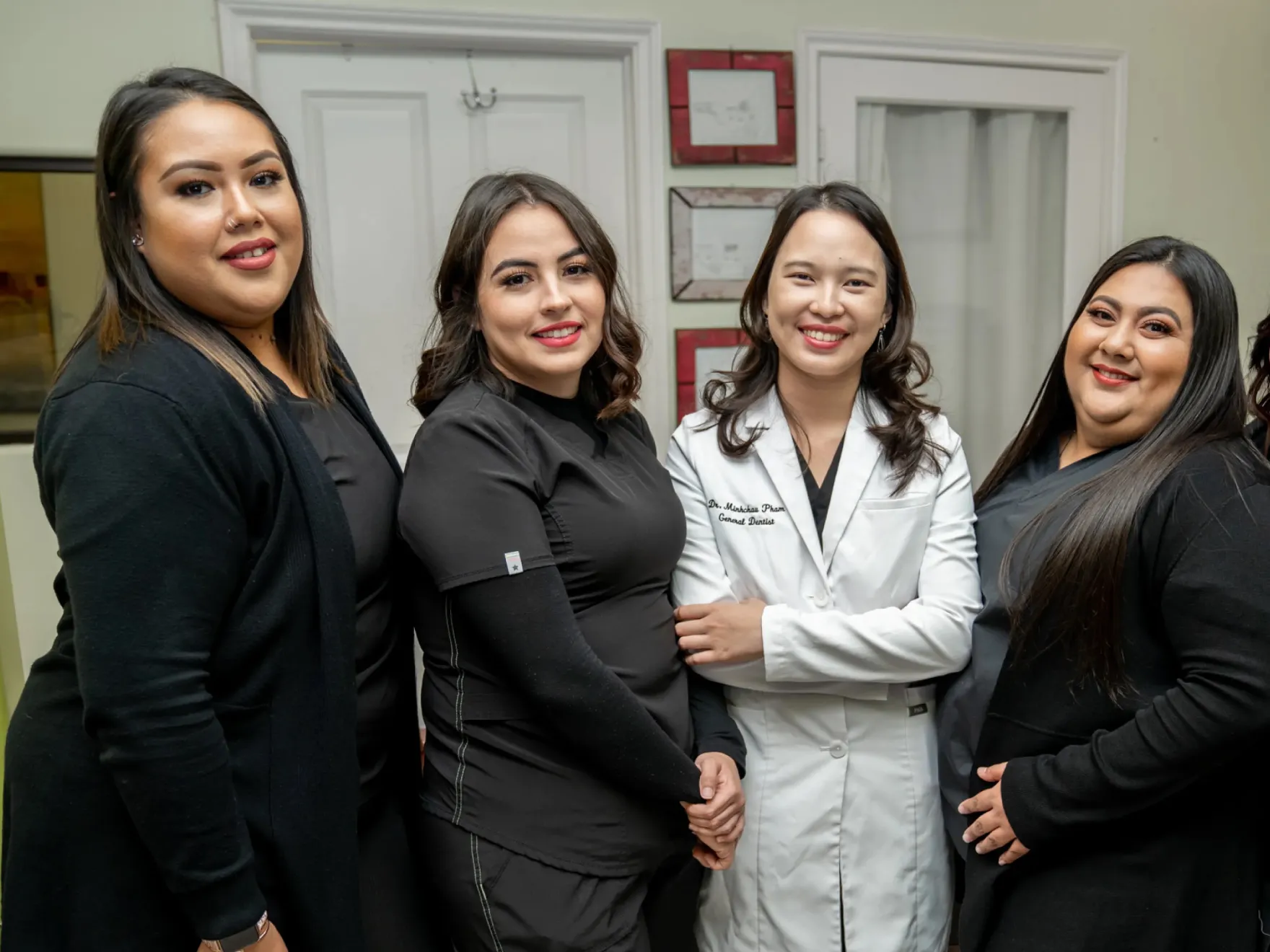 Four smiling women standing indoors, with one wearing a white lab coat labeled 'Dr. Minhcha Pham General Dentist' and the others in black clothing.