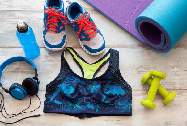 Flat lay of fitness gear including blue and white sneakers with red laces, floral sports bra, yellow dumbbells, blue headphones, blue water bottle, and rolled purple and blue yoga mats.