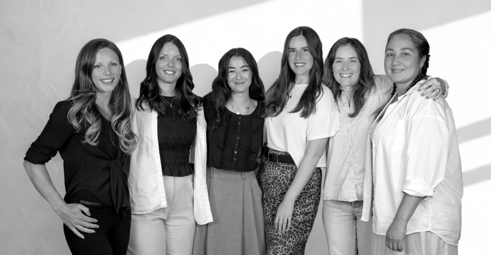Six women standing side by side against a plain white wall, smiling at the camera in a black and white photo.