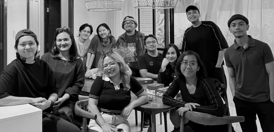 Group of eleven diverse young adults smiling and seated together in a modern room with hanging wicker lamps.