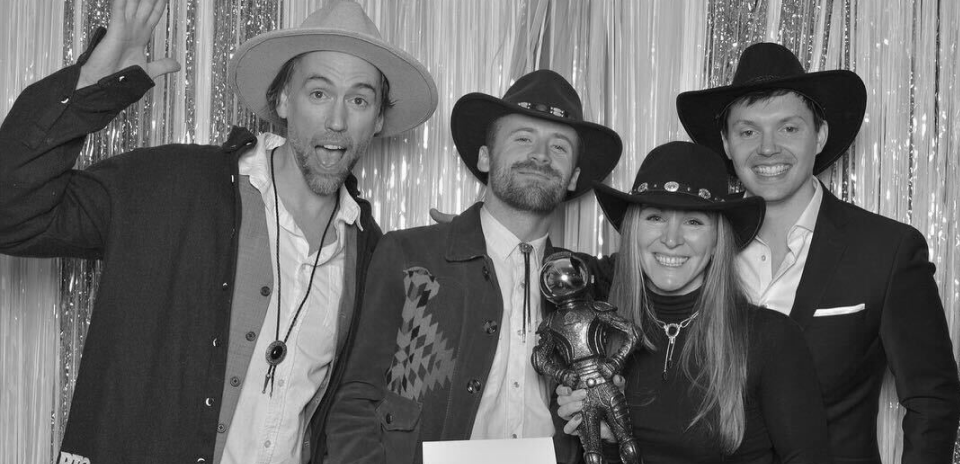 Four people wearing cowboy hats posing in front of a shiny backdrop, one holding an astronaut trophy.