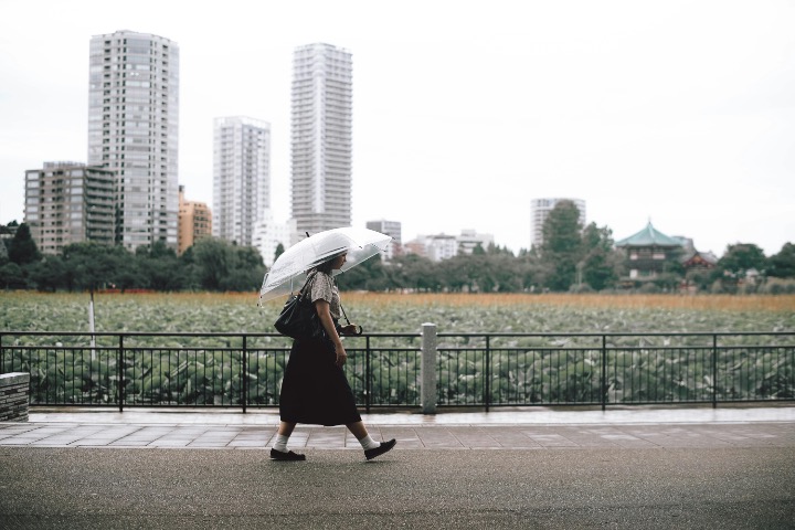 A person with a transparent umbrella walking along a riverside with high-rise buildings in the background in Tokyo