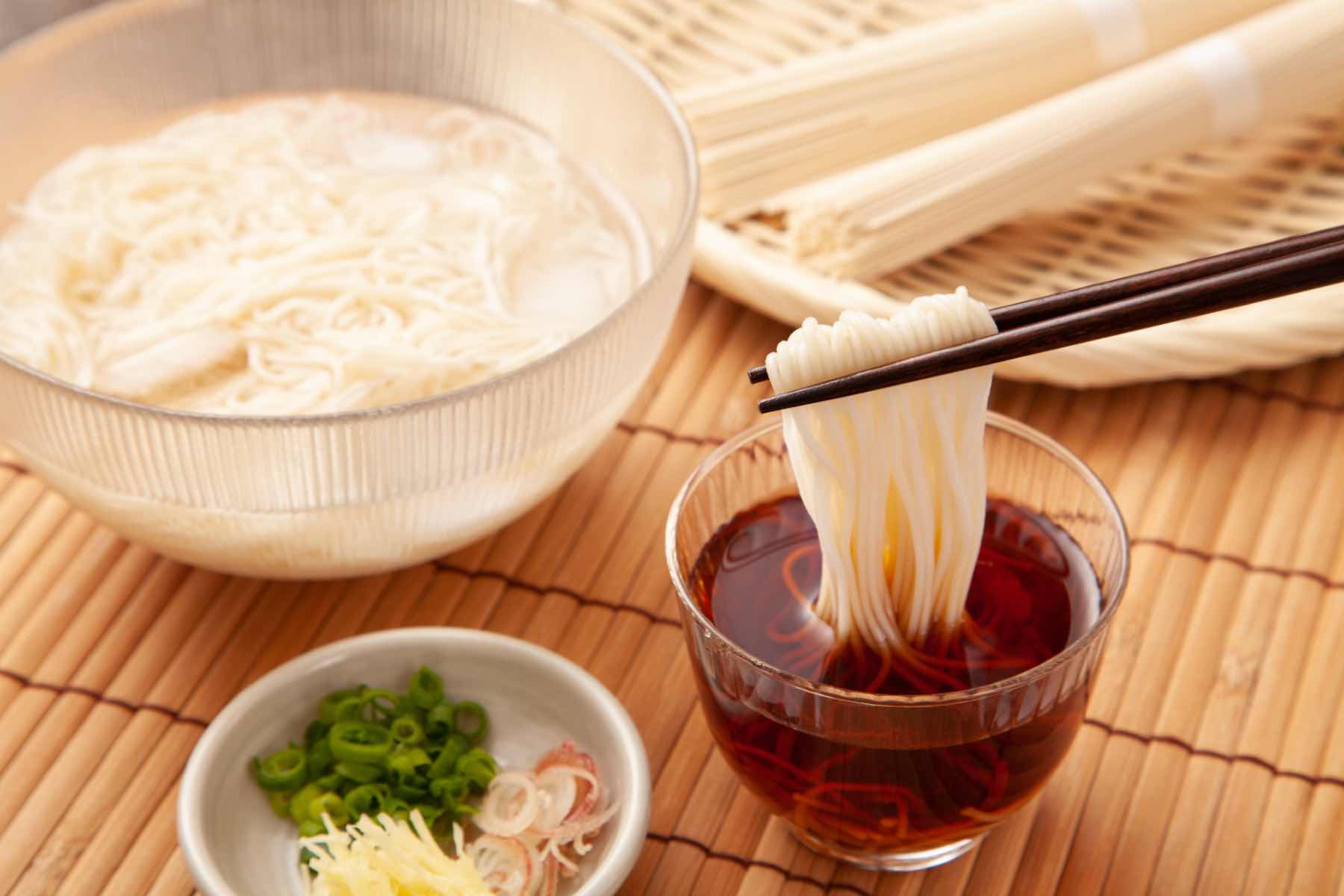 A bowl of chilled sōmen noodles with dipping sauce and fresh garnishes on a bamboo mat