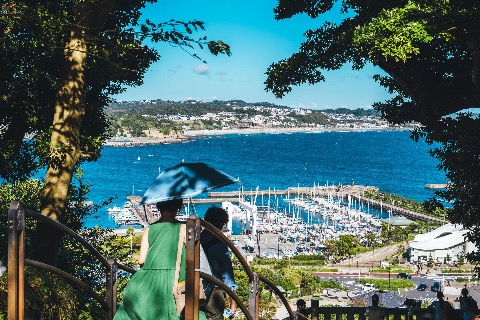 Two people with an umbrella descending stairs with a view of a marina and the ocean in Enoshima, Japan, on a bright summer day