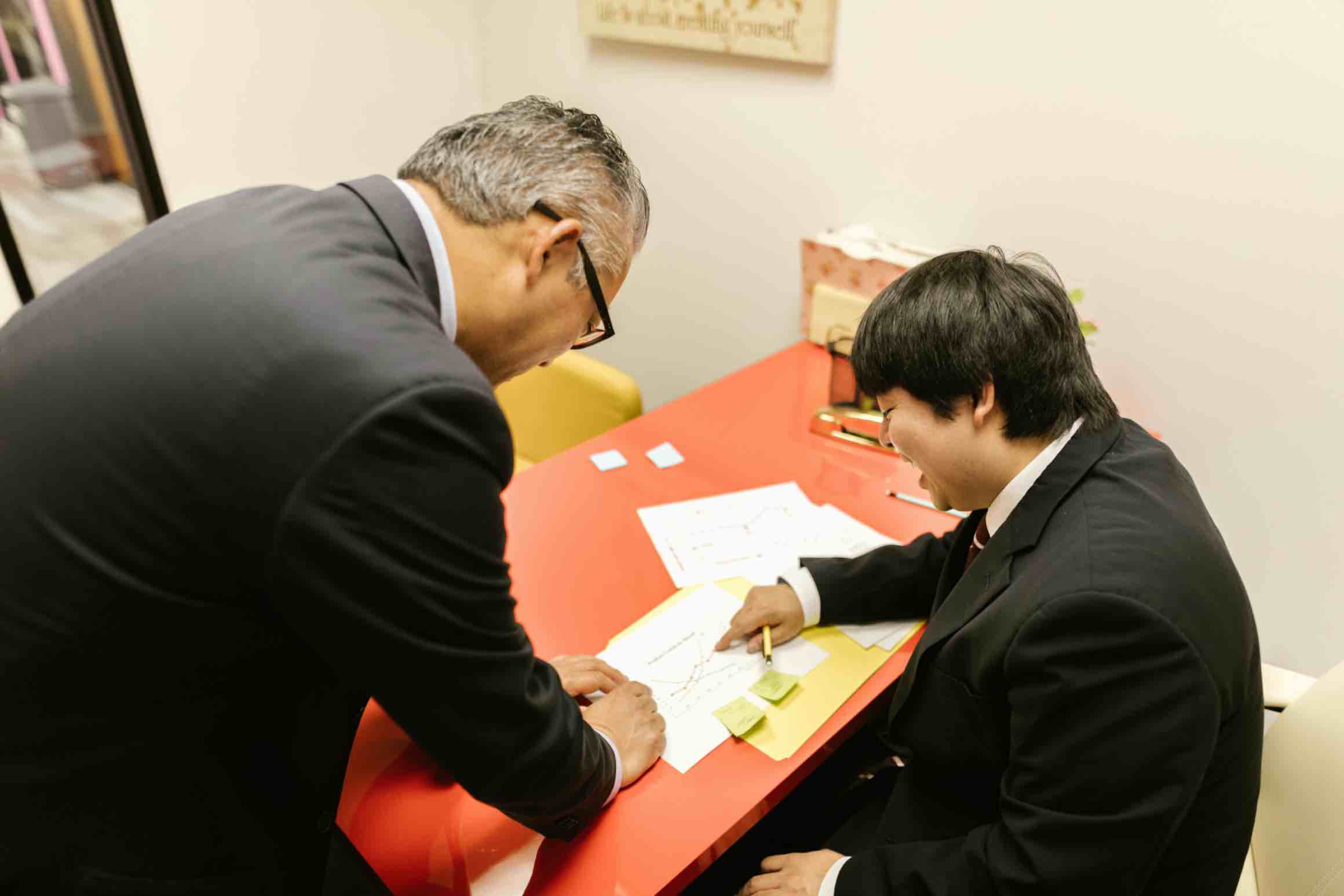 Two Japanese men in suits discussing documents at a red desk