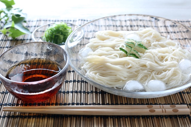 Bowl of chilled sōmen noodles with ice cubes and dipping sauce on a bamboo mat.