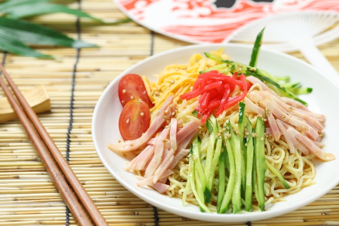 A colorful plate of hiyashi chūka topped with sliced cucumber, ham, egg, tomato, and red pickled ginger.