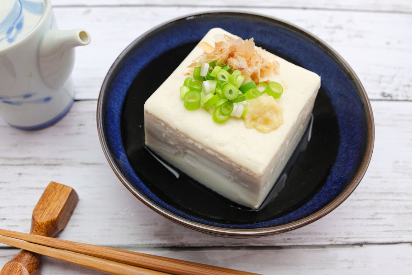 A chilled block of hiyayakko tofu topped with green onions, grated ginger, and bonito flakes, served in a black dish.