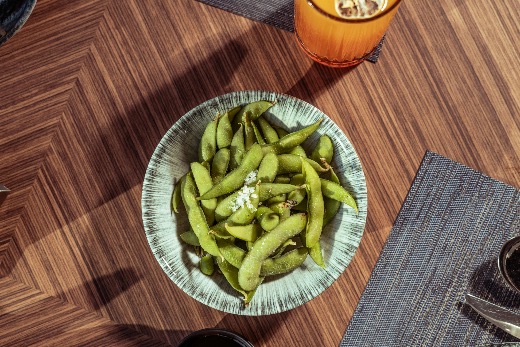 Bowl of salted edamame served with iced drinks on a wooden table.