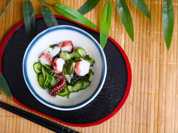 Bowl of sunomono with cucumber, wakame seaweed, and octopus slices on a bamboo tray.