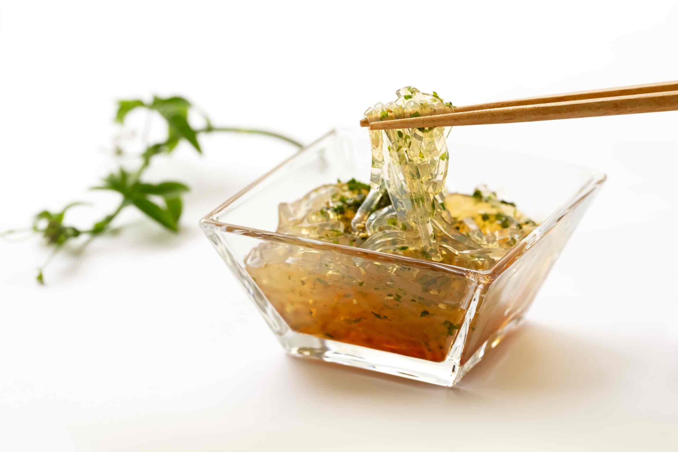 Clear tokoroten noodles being lifted with chopsticks from a glass bowl with vinegar sauce.