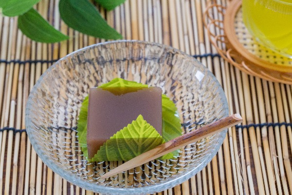 A glass bowl of mizuyōkan: a translucent red bean jelly served on a leaf, with a traditional dessert pick.