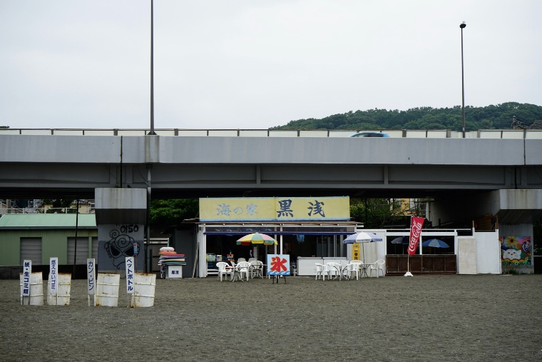 A small beachside food shack (umi no ie) with umbrellas and tables set up on the sand beneath an overpass.