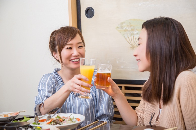 Two women smiling and clinking beer and juice glasses at a Japanese restaurant.