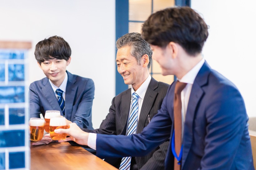 Three businessmen in suits clinking beer glasses at a counter in a modern Japanese bar.