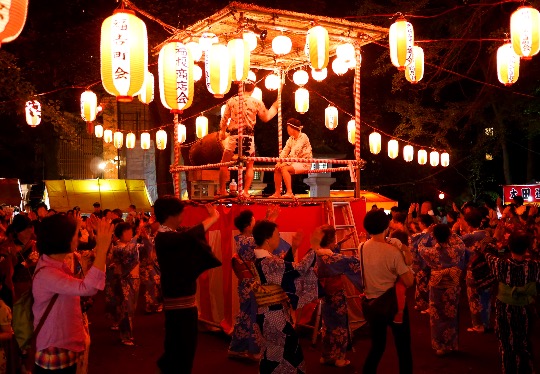 People in yukata dance around a yagura tower during a Bon Odori festival at night, surrounded by lanterns.