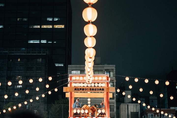 Lanterns lead toward a yagura tower where taiko drummers play during a nighttime Obon festival.