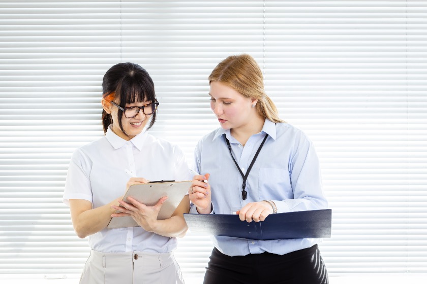 Two young interns, one Japanese and one international, reviewing notes on a clipboard in an office.