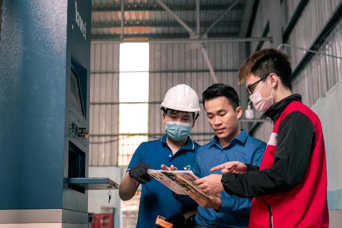 Engineers in a Japanese manufacturing setting reviewing technical documents, wearing safety equipment.