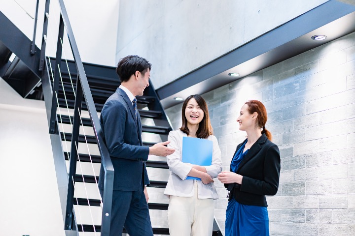 Three professionals, one Japanese woman, one foreign woman and one man, having a casual discussion in a modern Japanese office.