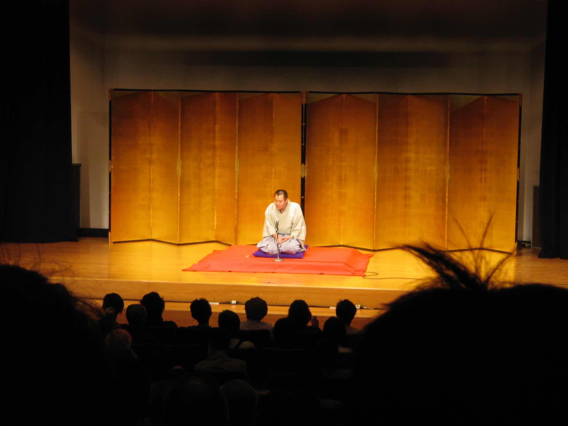 Rakugo performer kneeling on stage in front of a gold folding screen, performing for an audience.