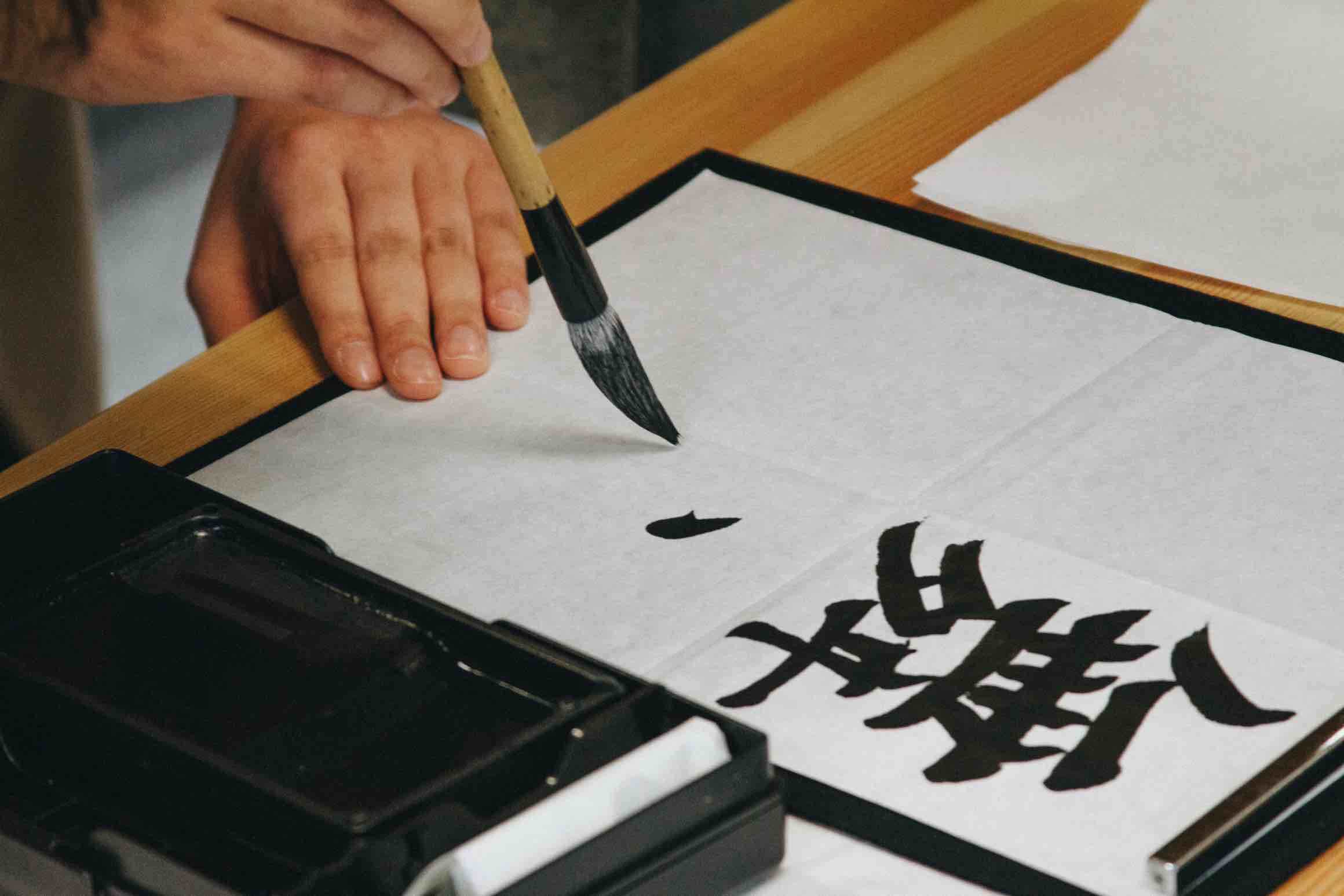 Japanese calligraphy in progress, brush writing black ink kanji on white paper.