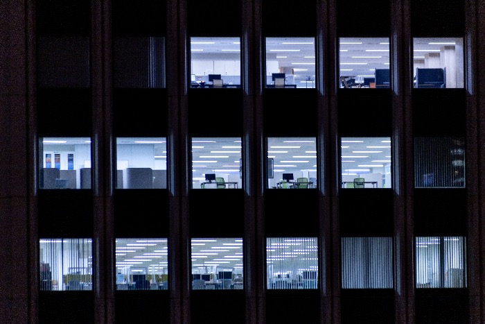 Corporate office interior with empty desks and bright overhead lighting visible through building windows at night.