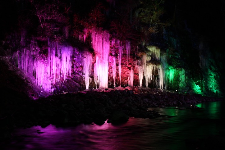 Large icicles illuminated at night along a rocky cliff beside a river in Chichibu, reflecting colored lights on the water.