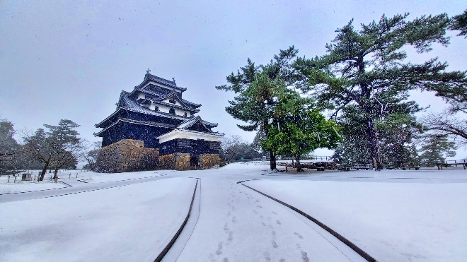 Matsue Castle standing on a stone foundation in winter, with falling snow and bare trees surrounding the grounds.