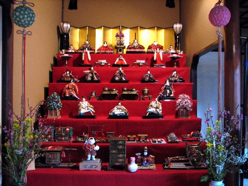 Multi-tiered Hinamatsuri doll display with emperor and empress dolls, attendants, and miniature court accessories arranged on red steps.