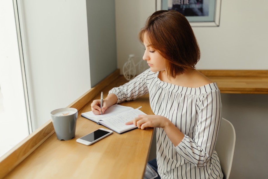 A woman sitting by a window writing in a notebook with a cup of coffee and a phone on the table.