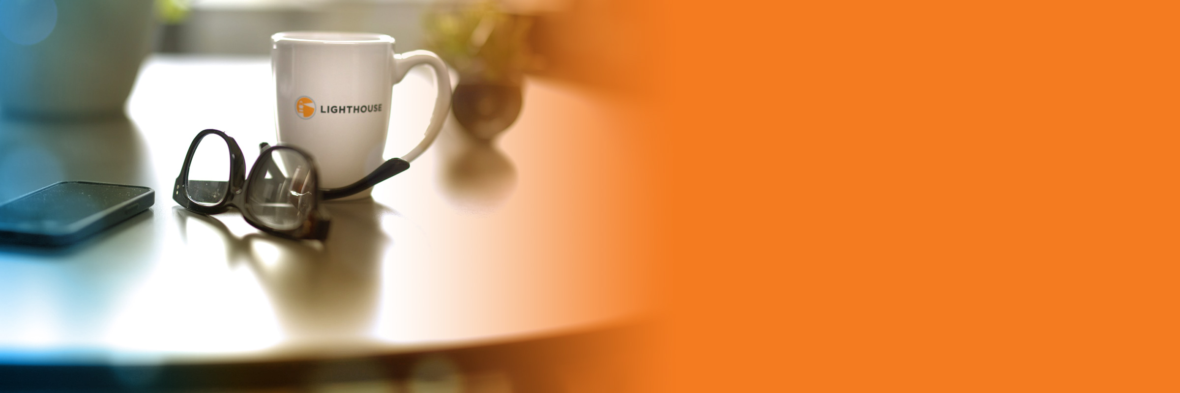 A Lighthouse coffee mug sits on a table with a pair of glasses and a phone.