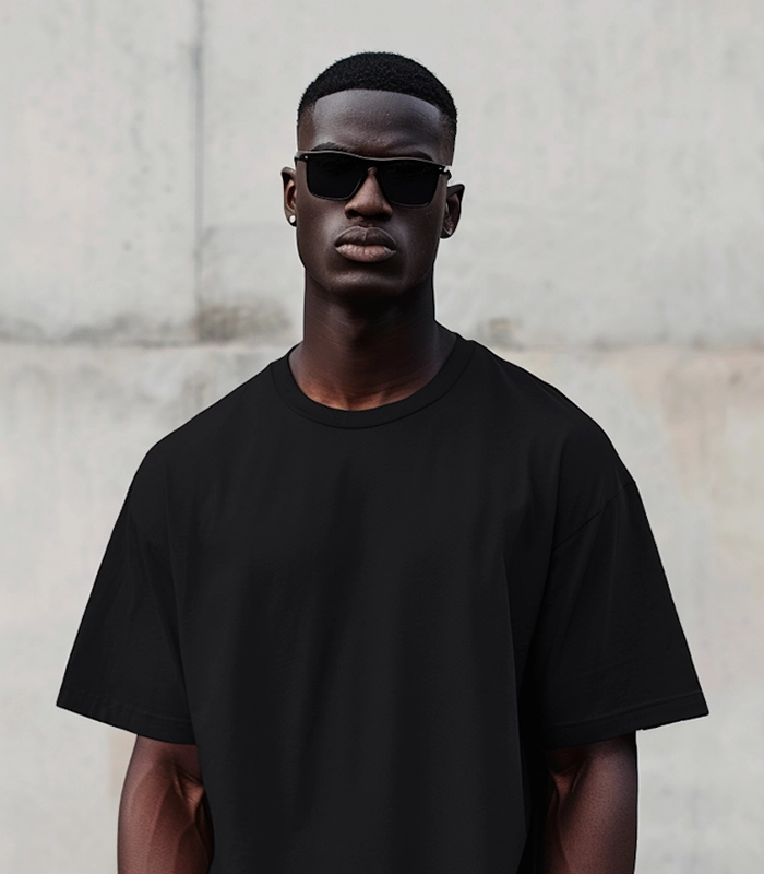 Man wearing black sunglasses and a plain black t-shirt standing against a light gray wall.
