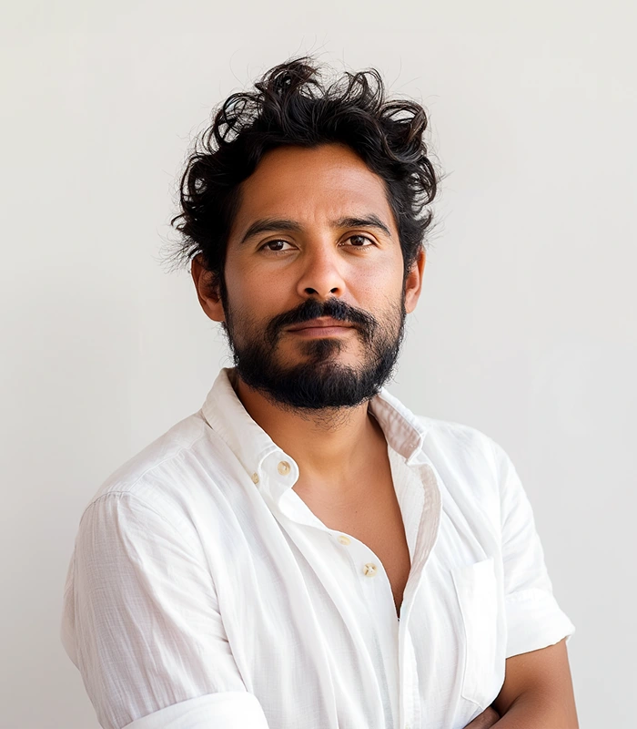 Man with curly black hair and beard wearing a white button-up shirt against a plain light background.