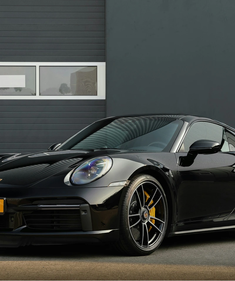 Front angled view of a sleek black sports car parked on a concrete surface against a dark wall with rectangular windows.