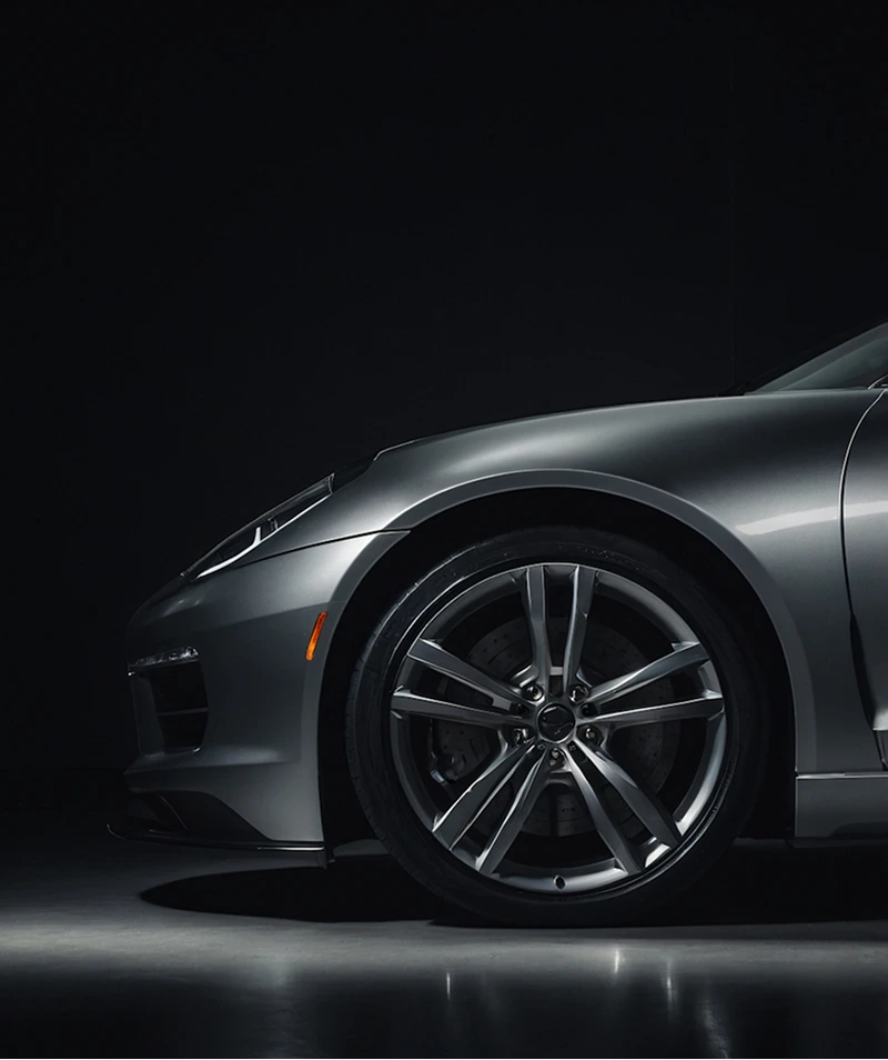 Close-up of the front left side of a silver sports car focusing on the wheel and fender against a dark background.