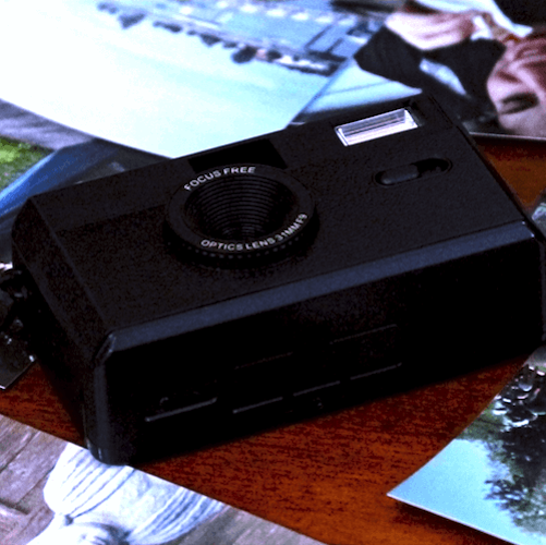 Black vintage point-and-shoot camera with focus free optics lens resting on a wooden surface surrounded by photographs.
