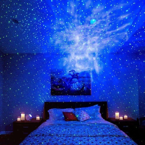 Bedroom with bed, pillows, and bedside tables lit by numerous candles and a ceiling projector casting blue and green star-like lights.