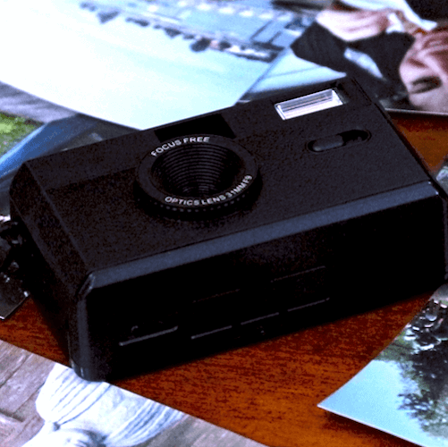 Black vintage film camera with a flash on a wooden surface surrounded by photographs.