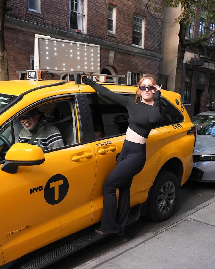 A woman in black crop top and pants poses while holding onto the roof of a yellow NYC taxi with a smiling driver inside.