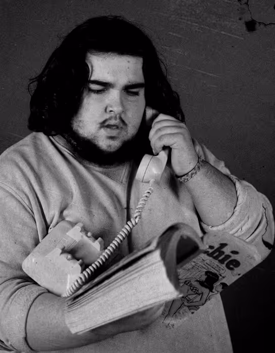 Man with dark curly hair and a beard holding a corded landline phone receiver to his ear with one hand and reading a book with the other.